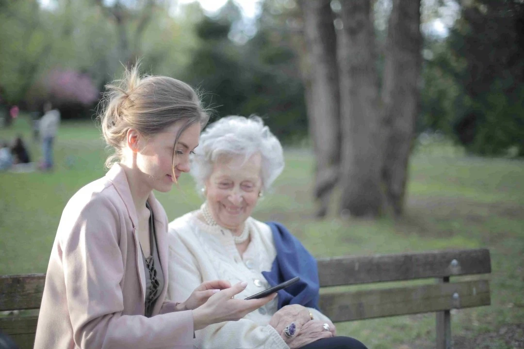Woman and elderly woman sitting on a park bench with a tree and green landscape in the background 