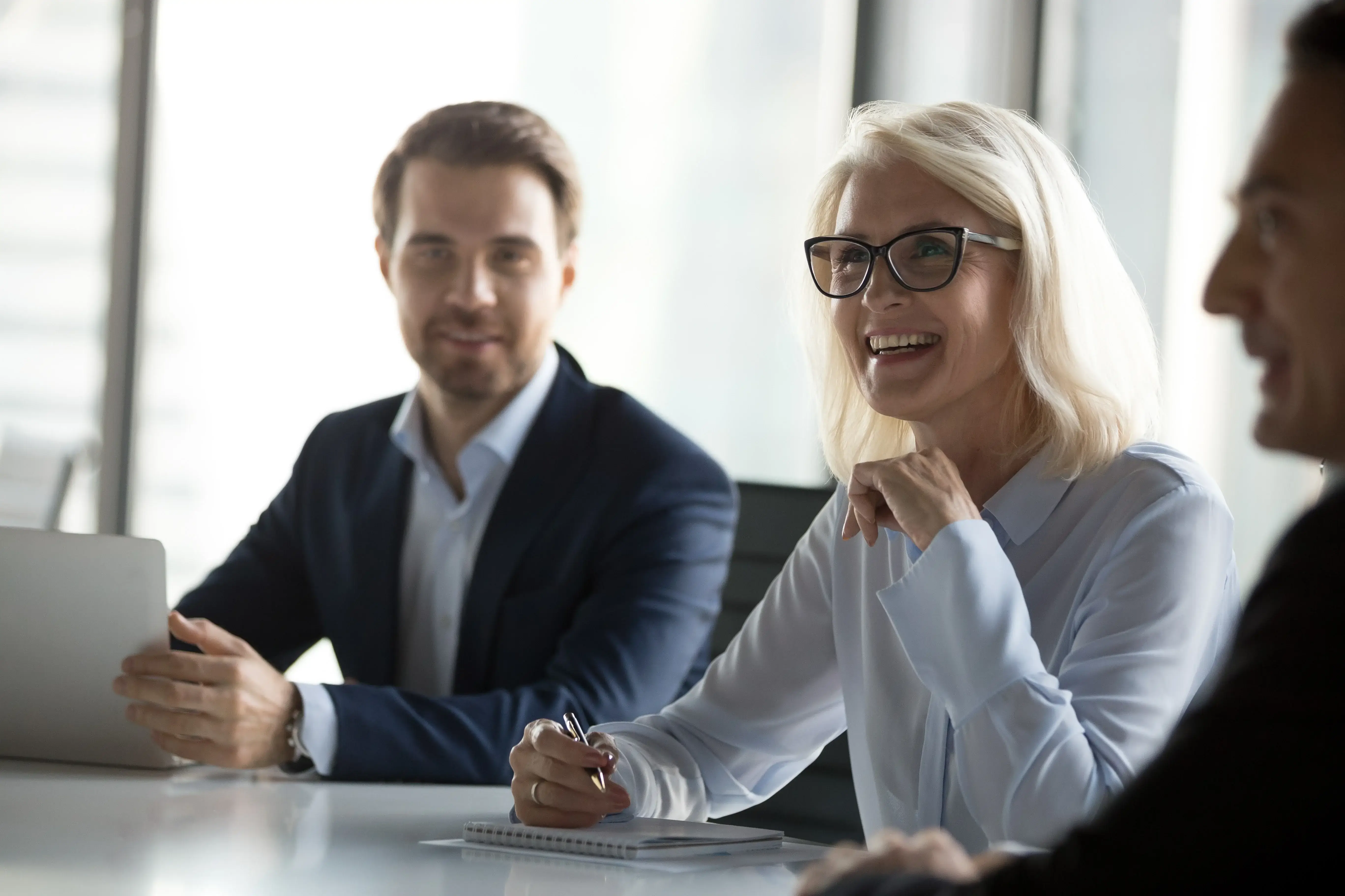 Smiling businesswoman with glasses listens and takes notes during a meeting, sitting at a conference table with two male colleagues. One man holds a laptop while they all engage in a positive and professional discussion.
