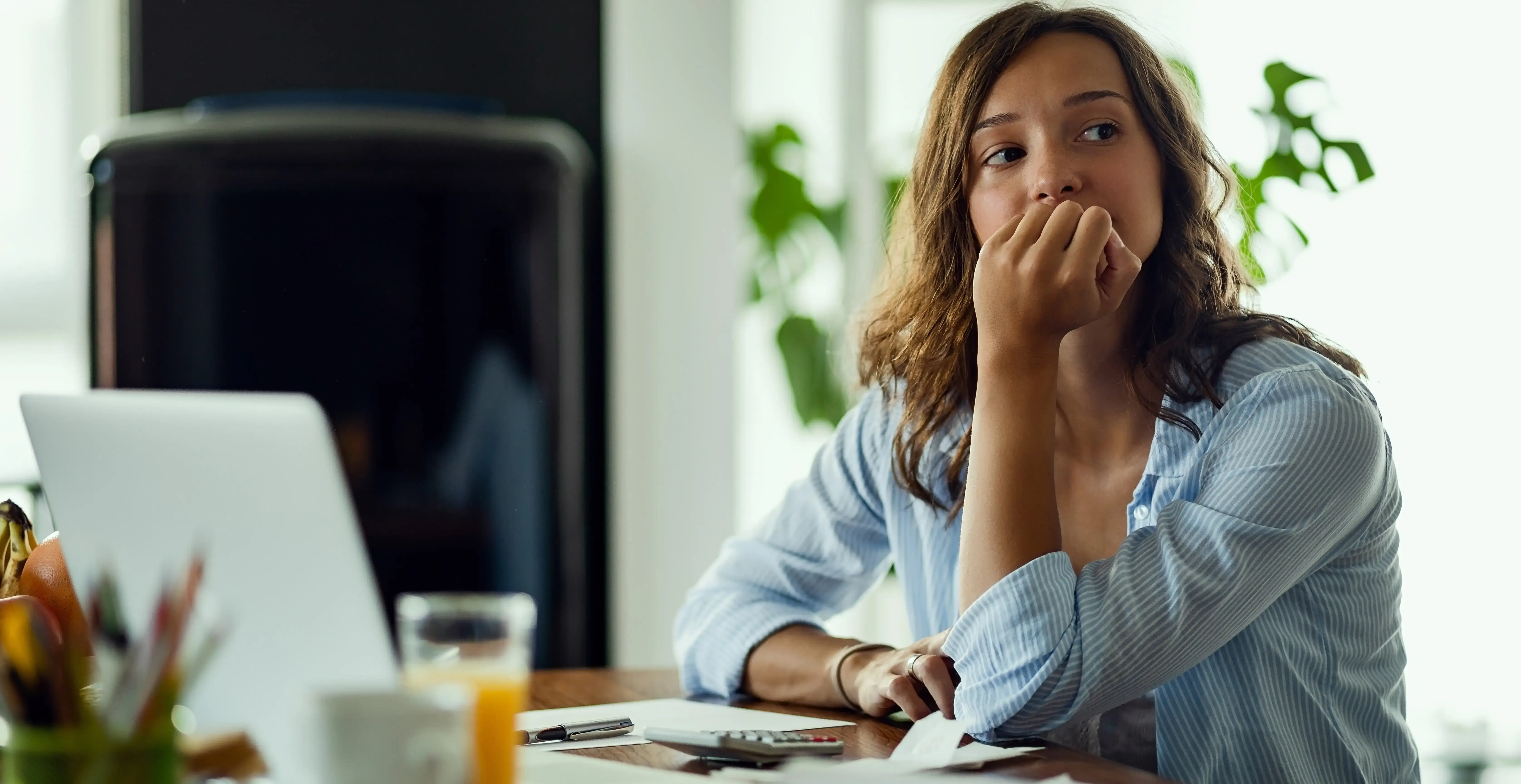 Thoughtful young woman sitting at a desk with a laptop, papers, and a glass of juice. She rests her hand on her face and looks away pensively, suggesting she is deep in thought or concerned while working from home.