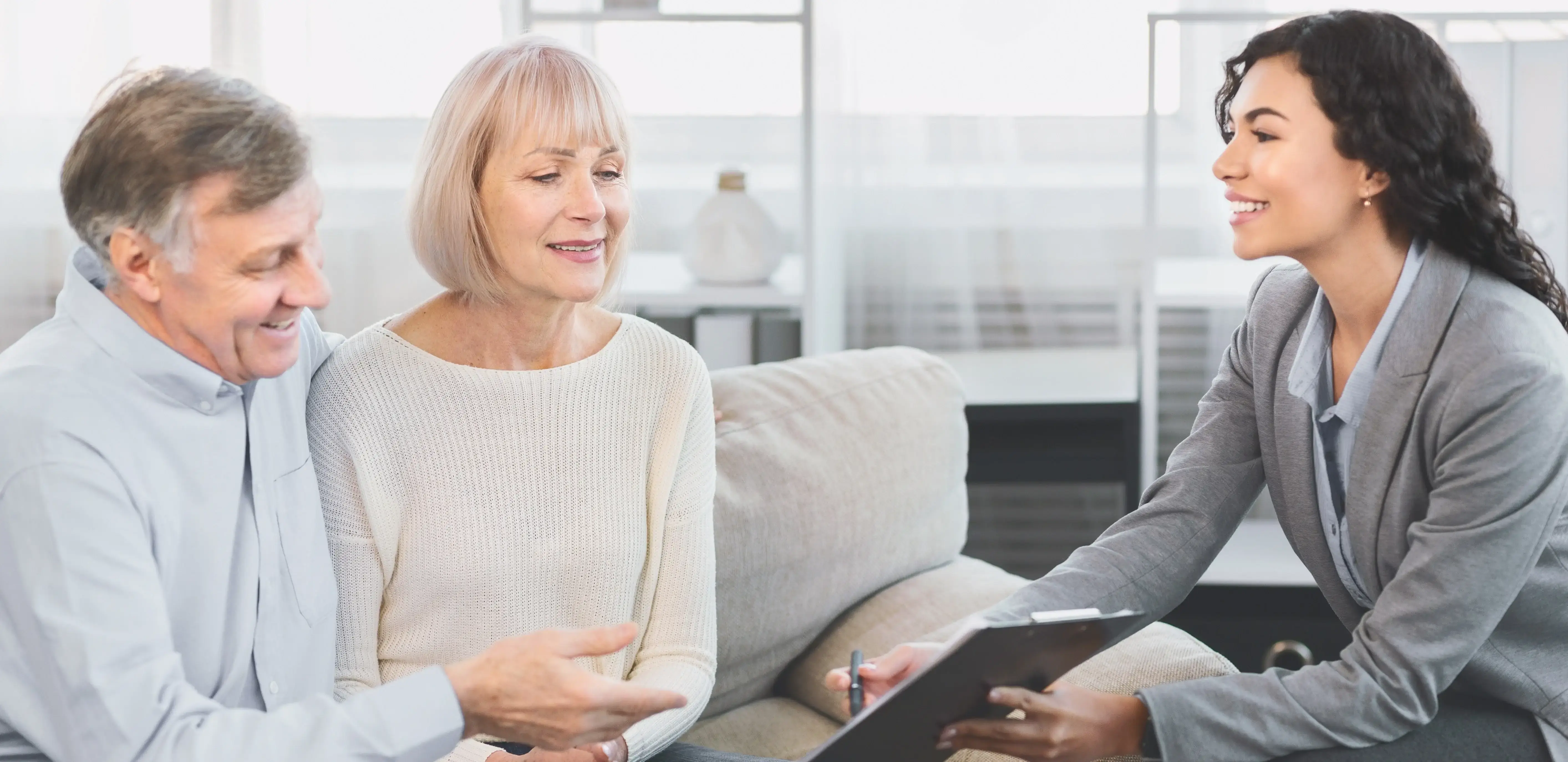 Smiling financial advisor meeting with an older couple at home, reviewing documents together on a clipboard.