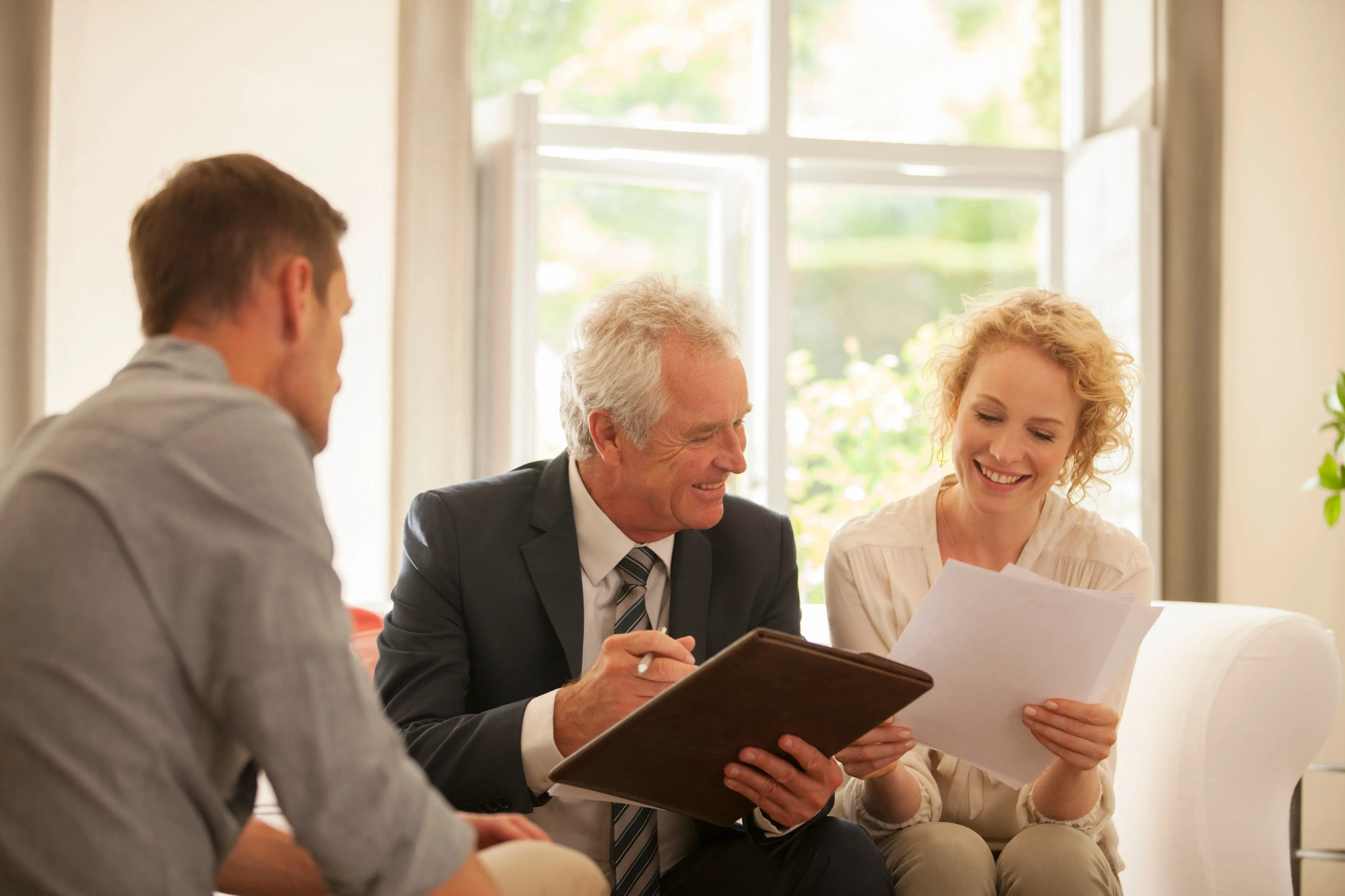 Smiling financial advisor in a suit holding a clipboard talks with a couple at home. The man listens attentively while the woman holds paperwork, and they all look engaged and positive during the discussion