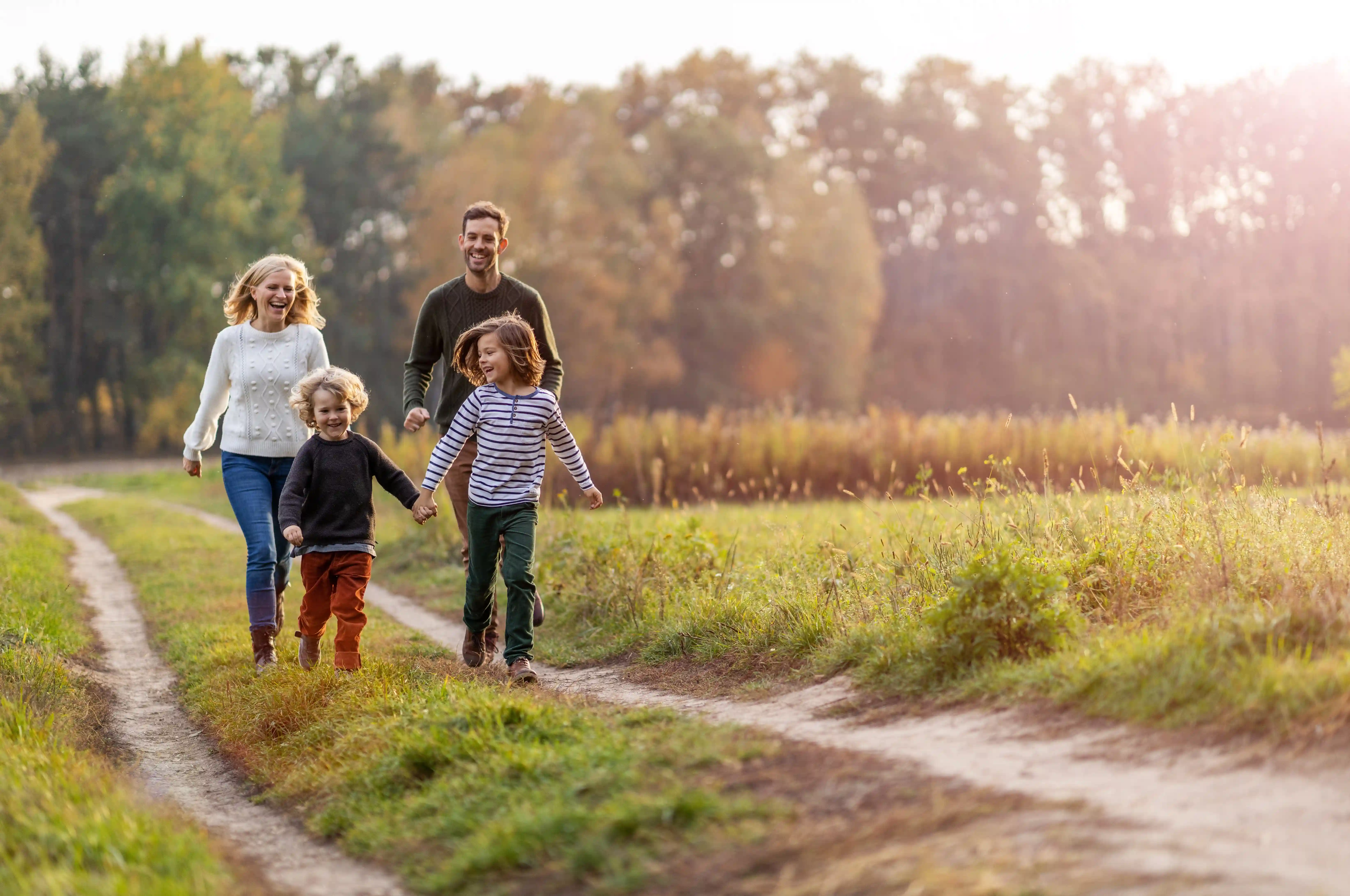 Happy family of four walking outdoors on a sunny day. Parents follow behind as two children hold hands and run along a grassy path, surrounded by fields and trees in the background.