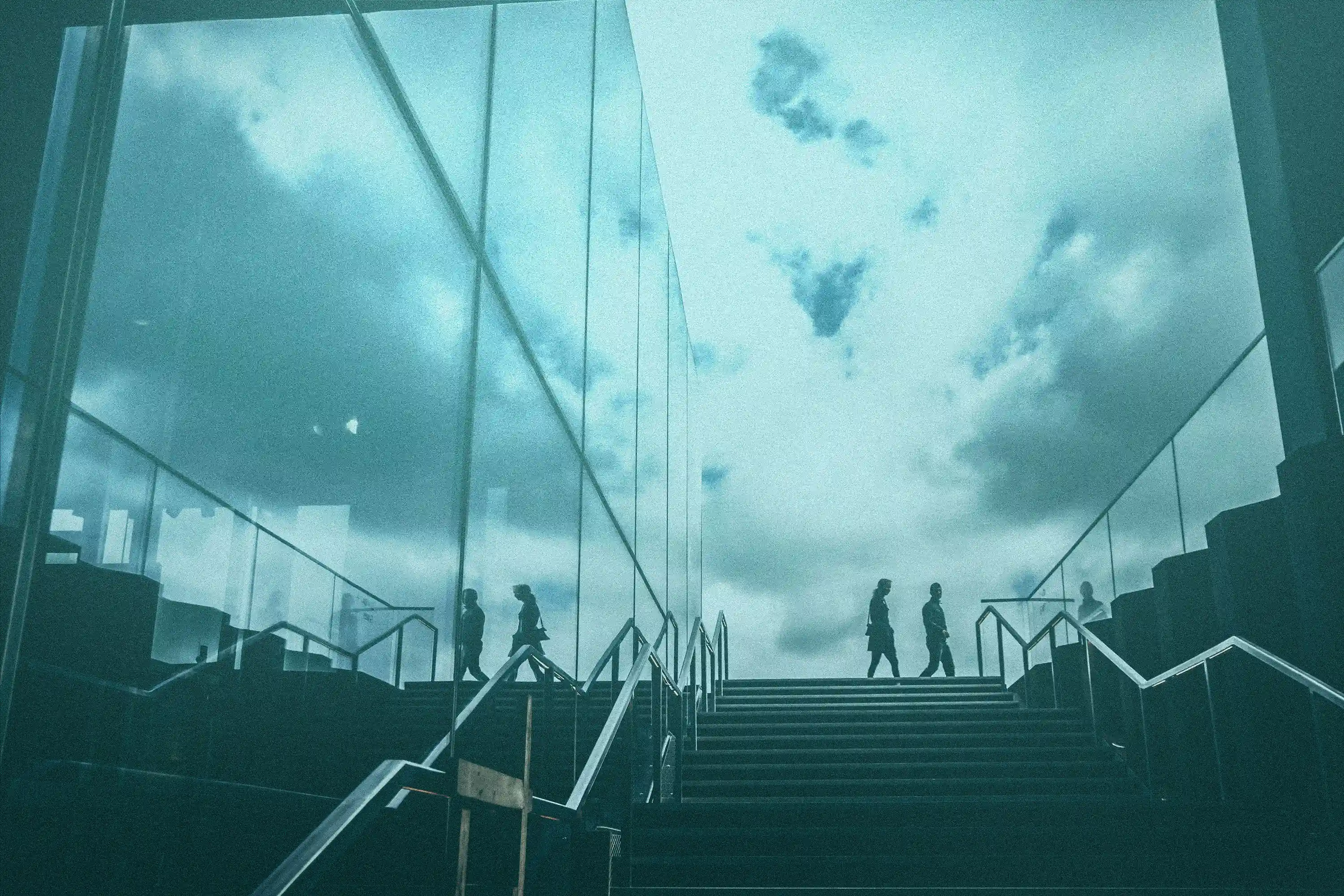 A modern glass-walled staircase leading upward, with several silhouetted people walking along the top landing. The reflective surfaces mirror the cloudy sky above, creating a symmetrical, teal-toned urban scene with a minimalist, slightly surreal atmosphere.