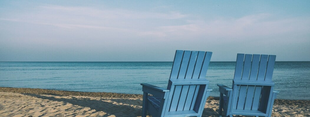 Two blue wooden chairs on a sandy beach facing the calm ocean under a clear sky.