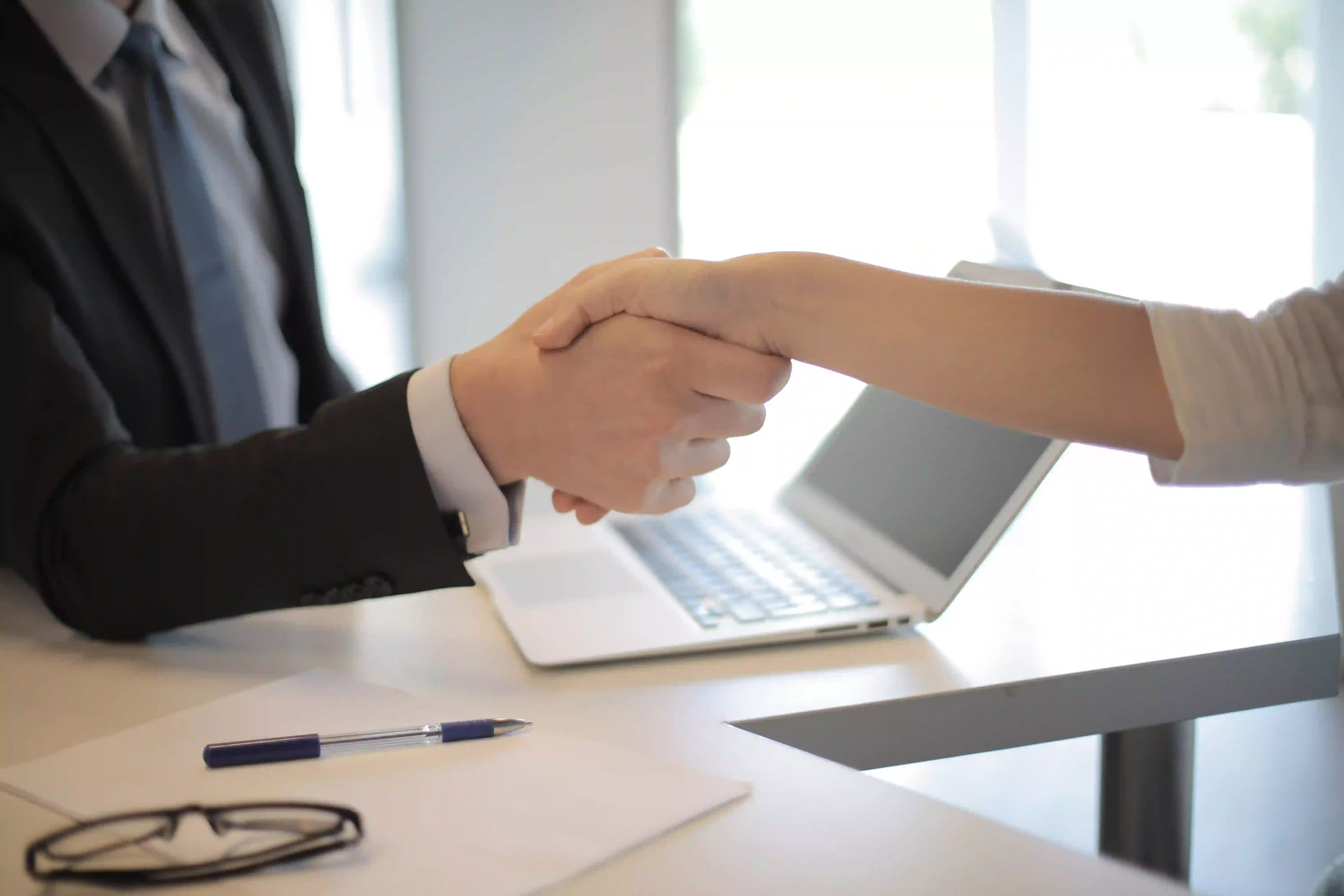 Businessperson and client shaking hands across a desk with a laptop, pen, and papers in view.