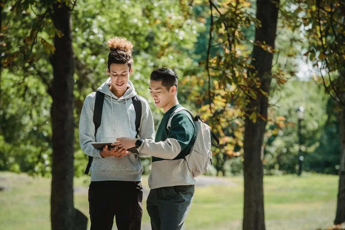 Two young men with backpacks standing outdoors in a park, smiling and looking at a tablet together.