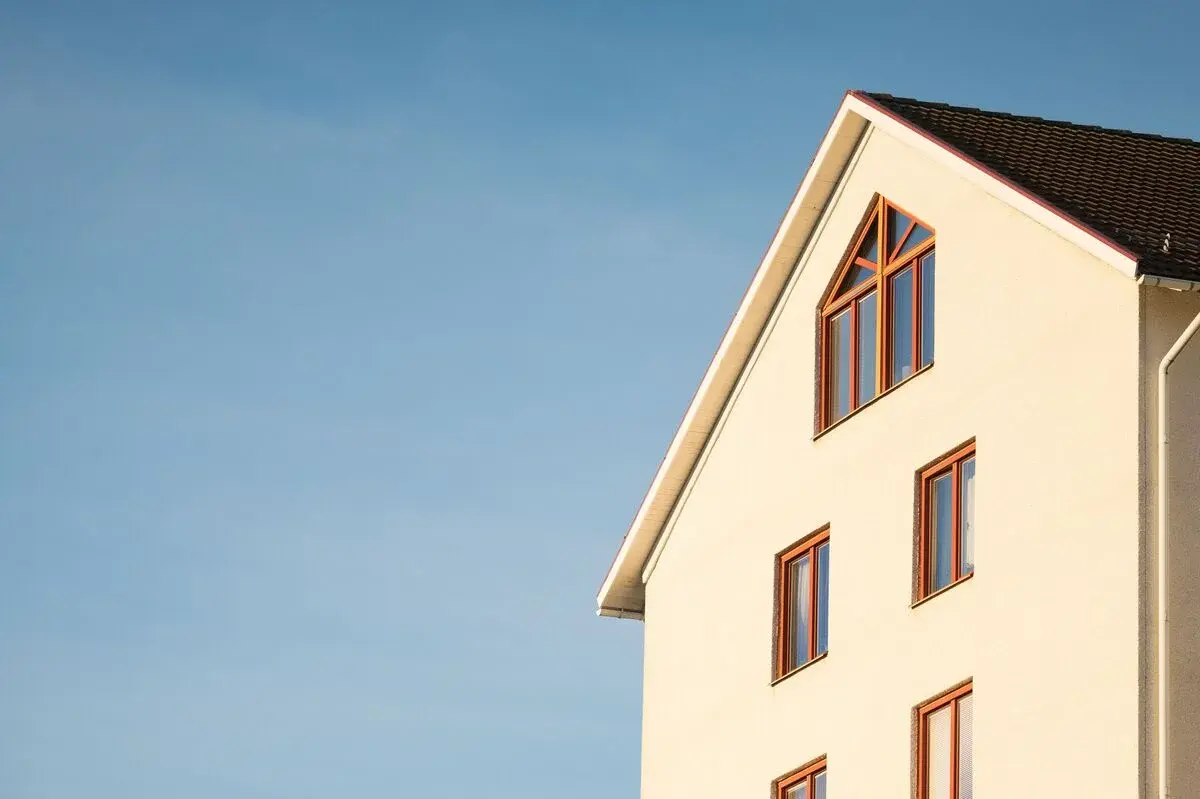 Close-up of the upper part of a beige house with triangular roof and multiple windows against a clear blue sky.