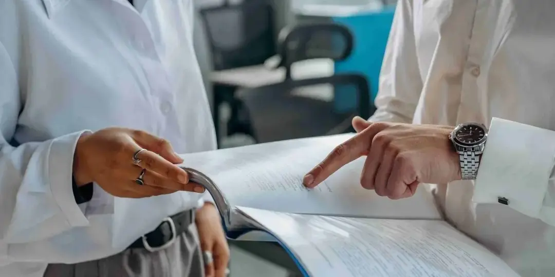 Two people in business attire are reviewing a document together in an office. One person is holding the open document while the other points at a section on the page.