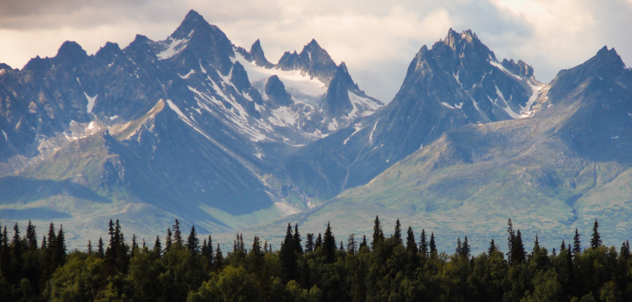 Scenic view of rugged snow-capped mountains with forested trees in the foreground under a partly cloudy sky.