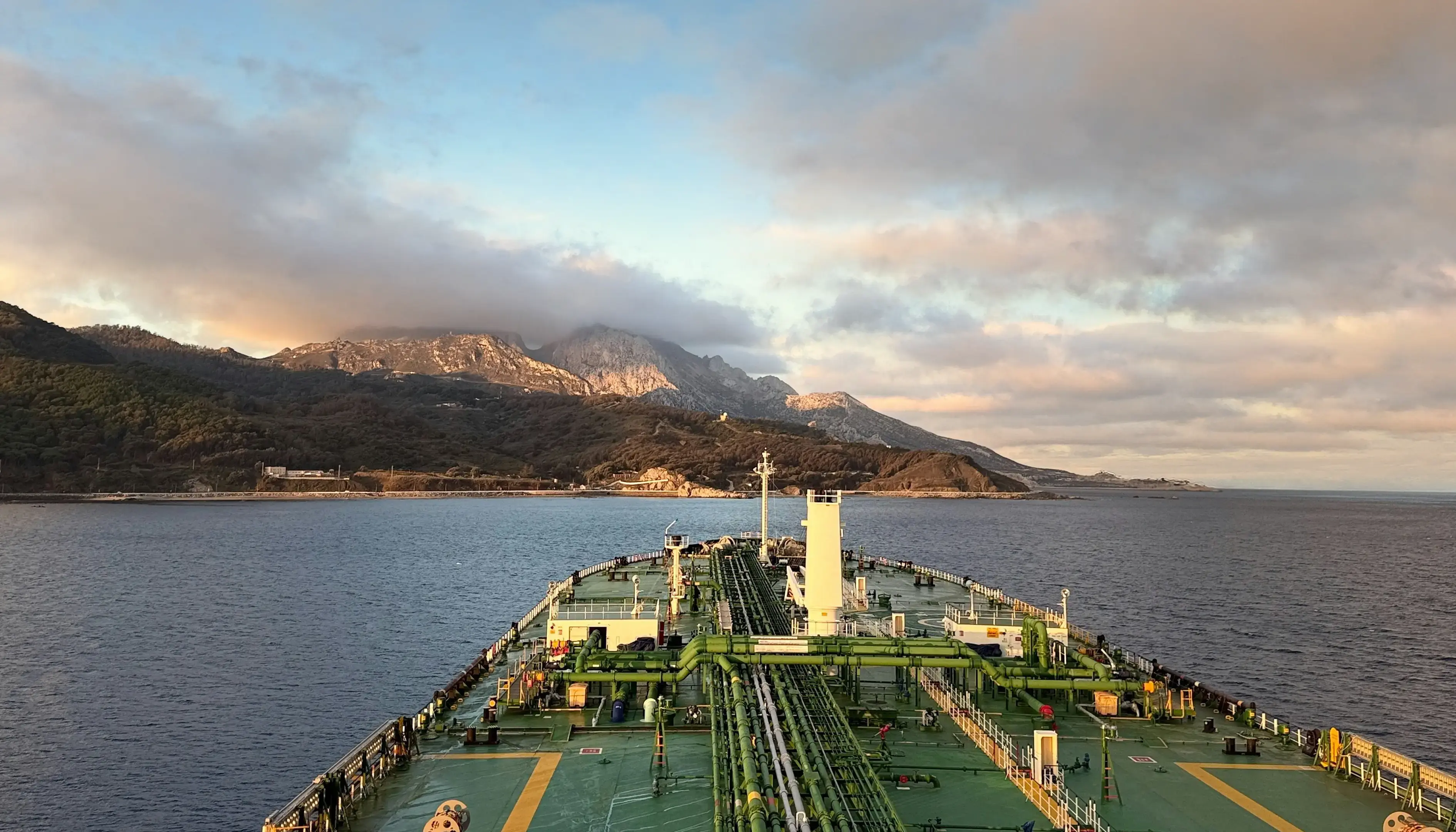 View from the deck of a large tanker ship with green piping and equipment, sailing on calm water toward a mountainous coastline under a sky filled with thick, dramatic clouds lit by soft golden light.