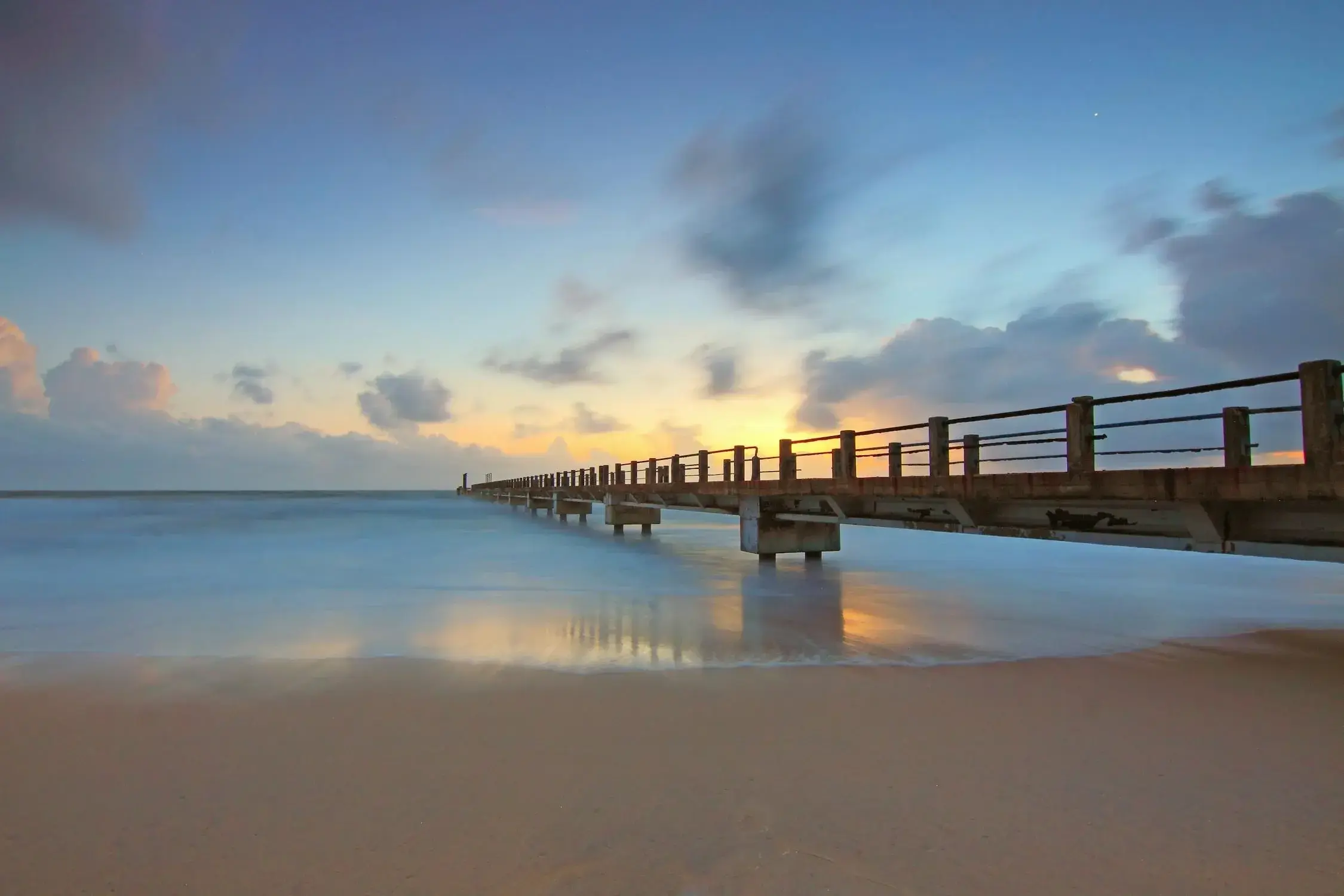 A long pier extending into the calm ocean at sunrise, with soft waves washing onto the sandy shore and clouds scattered across the colorful sky.