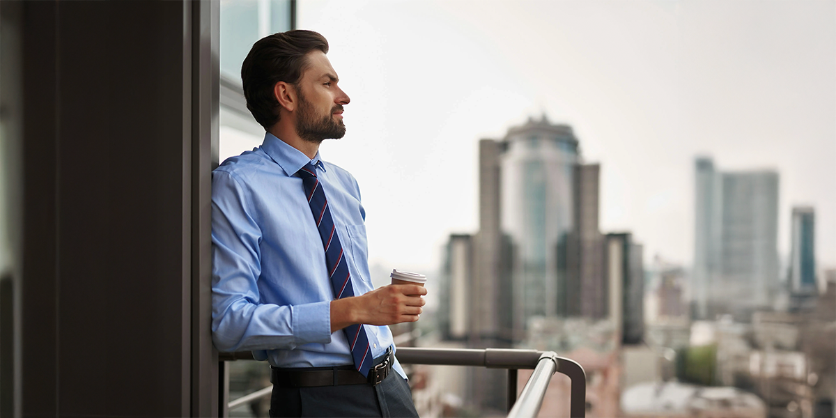 A well-dressed man with a beard stands on a balcony in a modern office building, holding a takeaway coffee and looking out over a city skyline with tall buildings in the background.