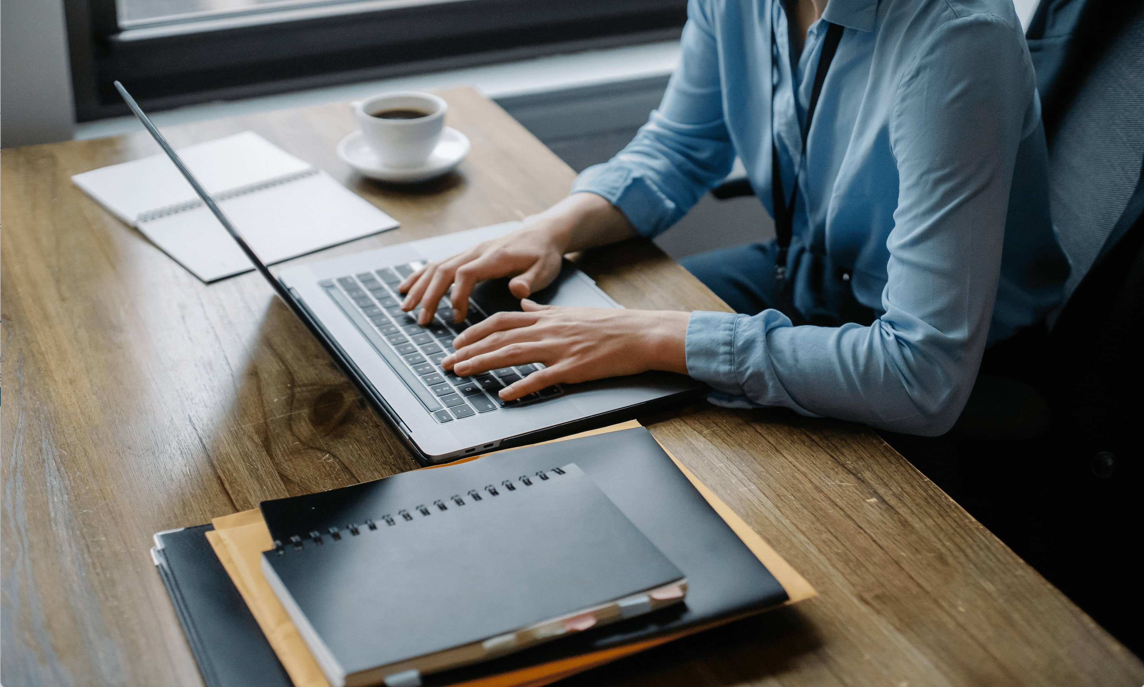 Professional working at a desk on a laptop, with notebooks, folders, and a cup of coffee nearby. The person wears a blue shirt and types on the keyboard, suggesting focus and productivity in an office setting.