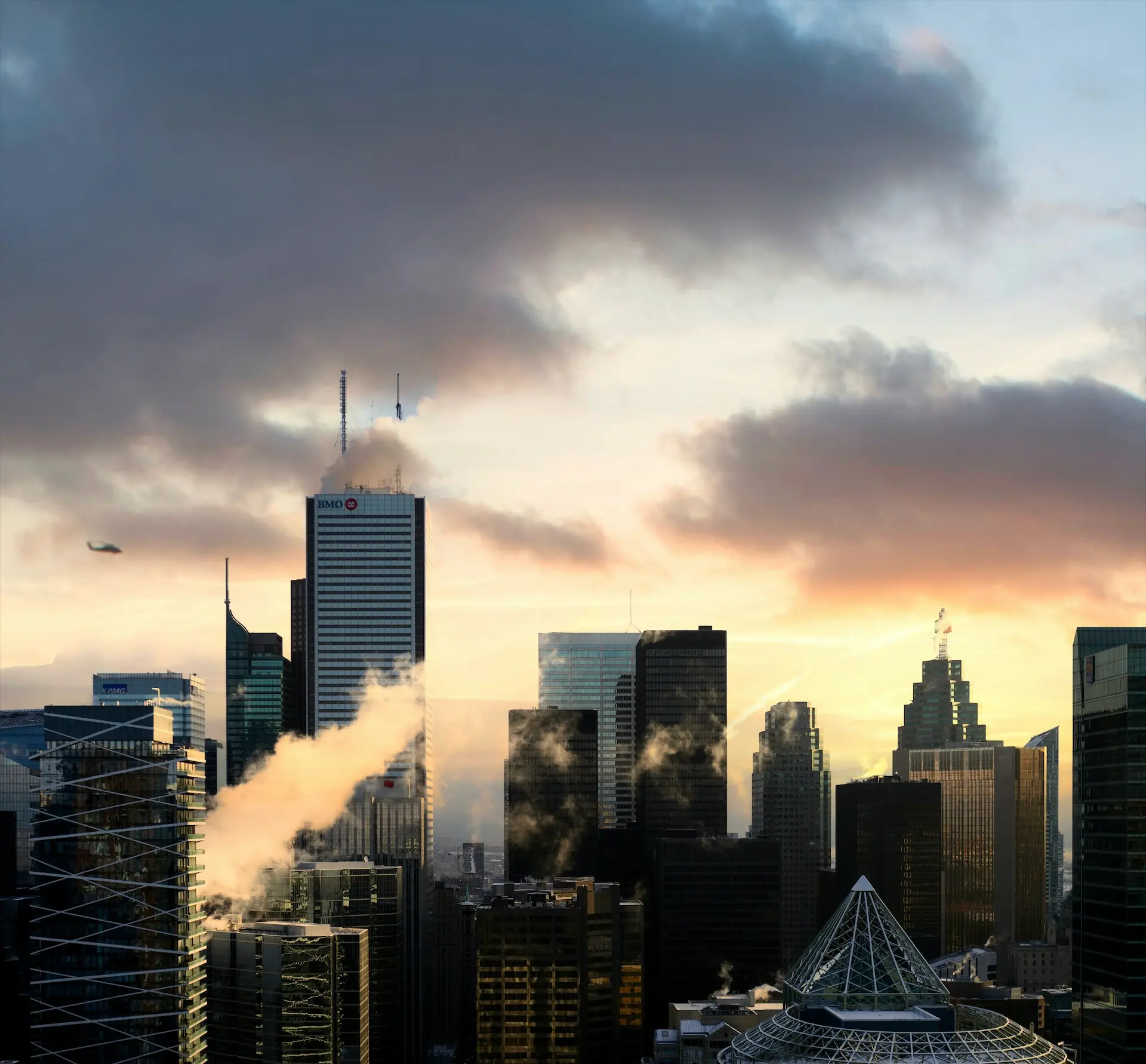A dramatic city (Toronto) skyline glows under a cloudy sunrise, with steam rising between skyscrapers and a helicopter crossing the sky.