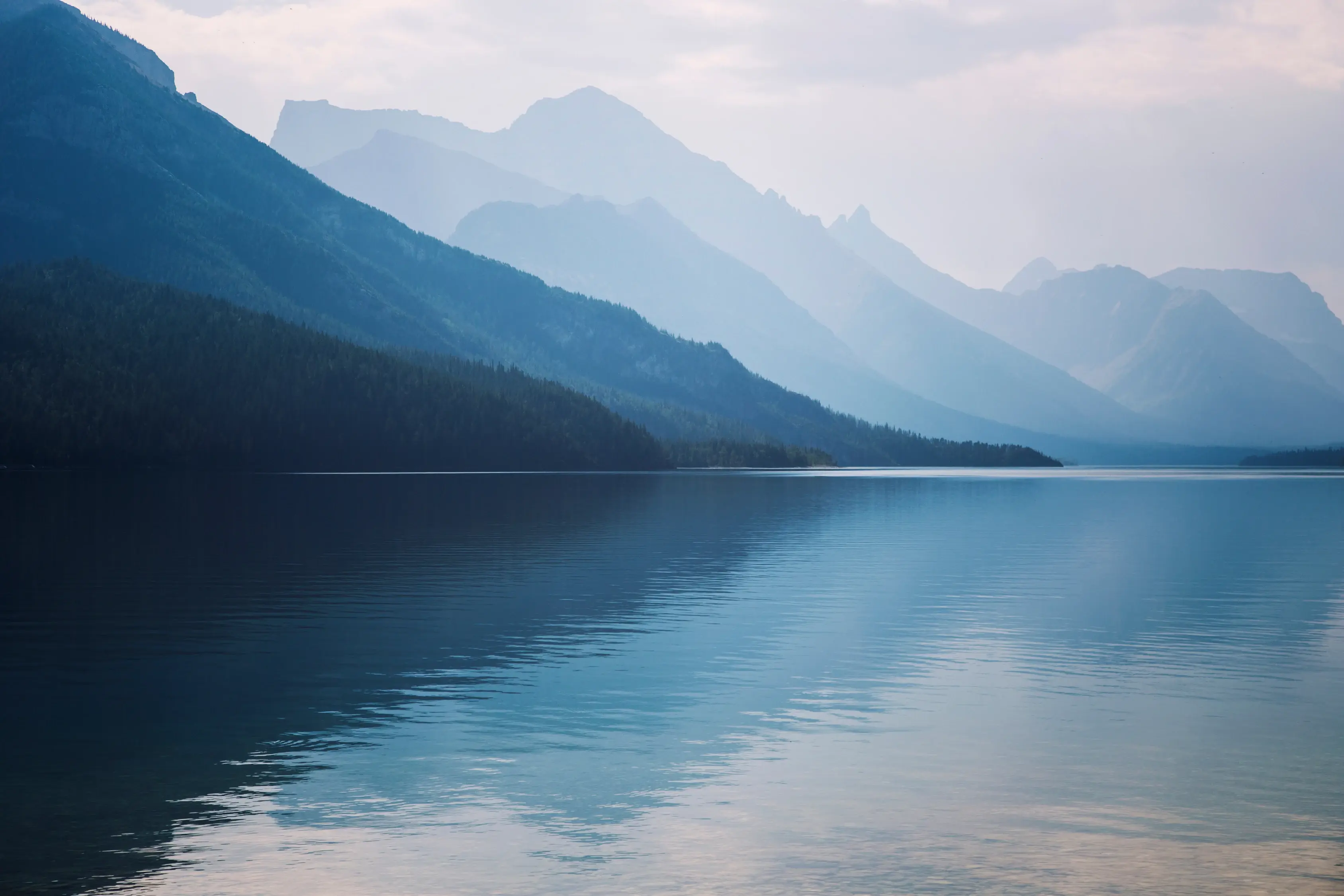 Serene lake with calm rippling water, surrounded by forested slopes and layered blue mountain ranges fading into the misty horizon