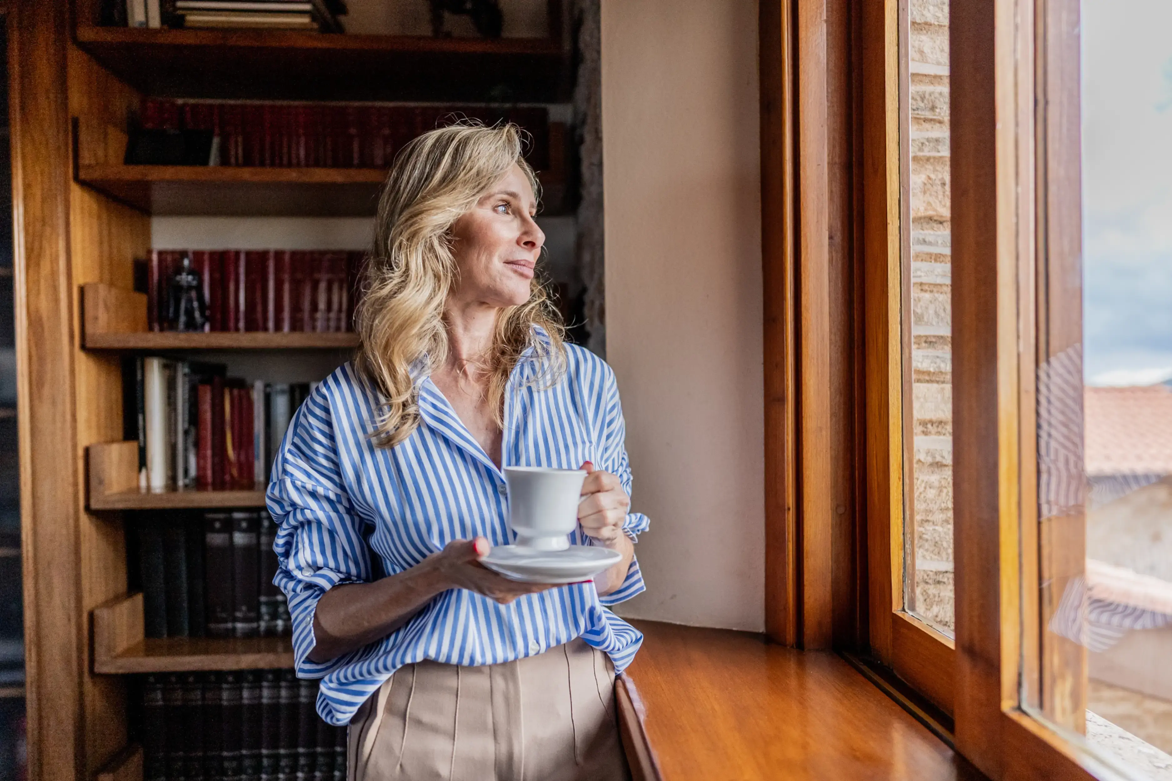 A woman with blonde hair stands by a window, holding a cup and saucer, wearing a blue-and-white striped shirt and beige pants, with bookshelves behind her.