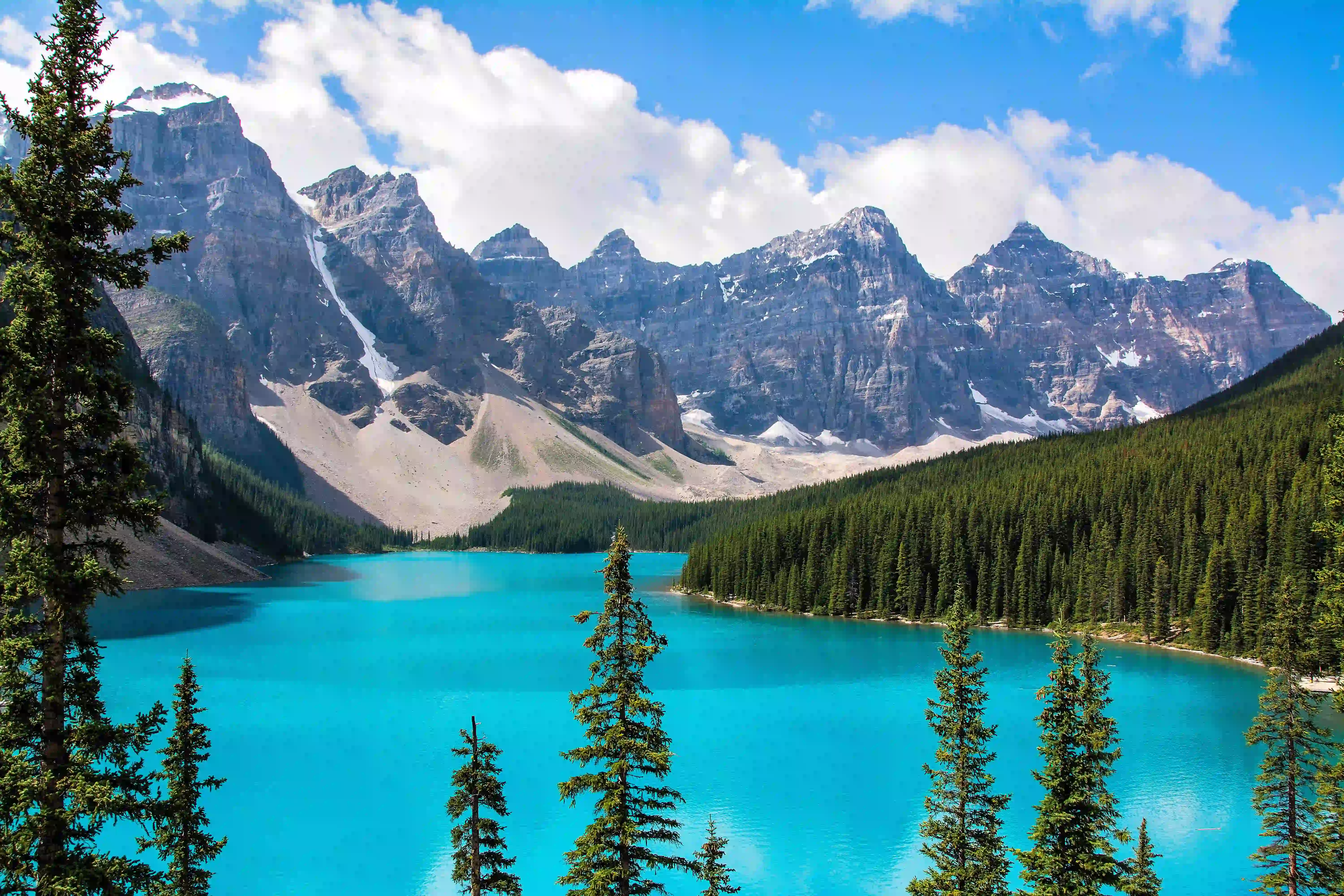 Turquoise lake surrounded by dense pine forests and rugged snow-capped mountains under a bright blue sky with scattered clouds.