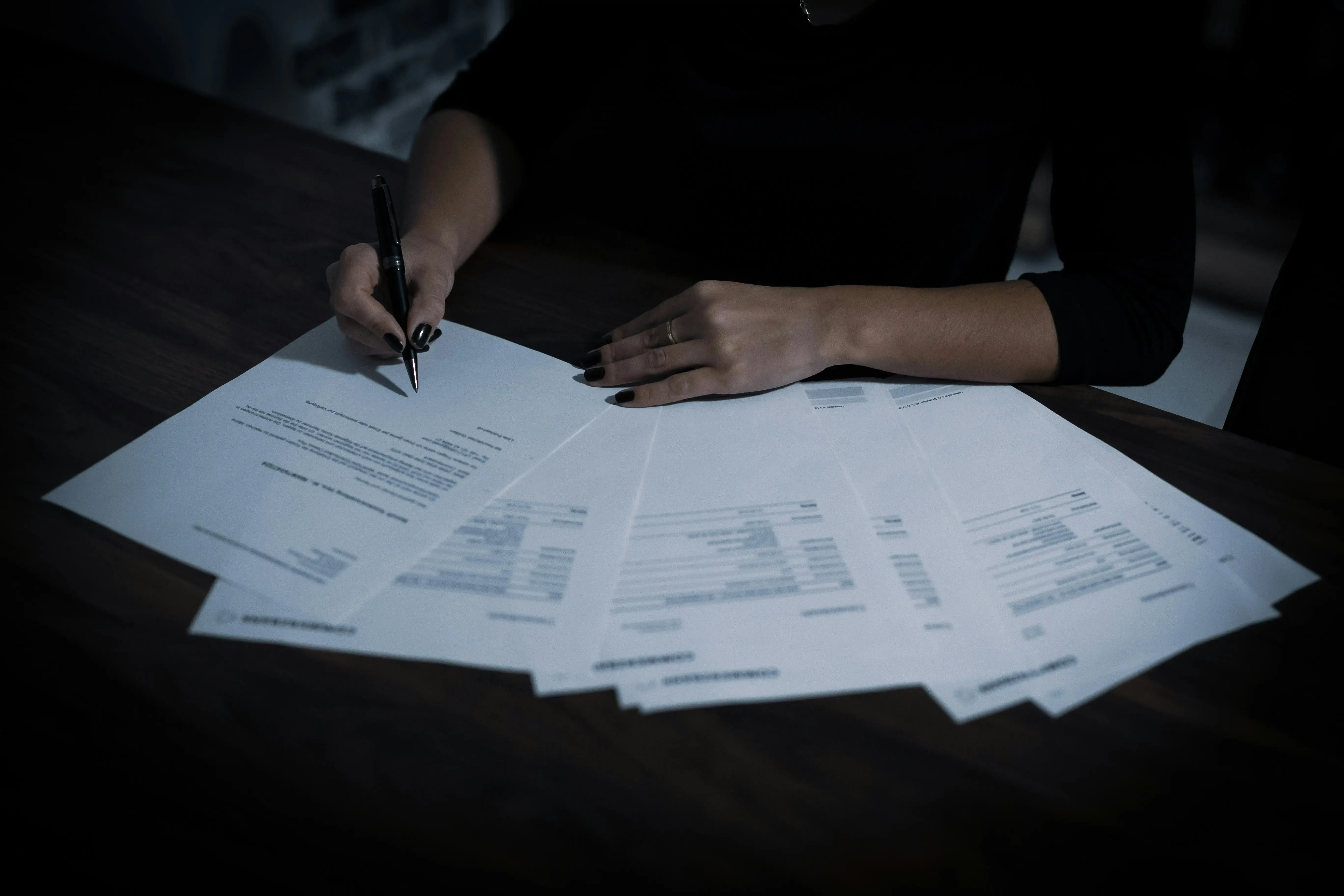 A person seated at a desk signs a document while several printed financial or formal papers are spread out in front of them. Only their hands and arms are visible, lit by dim, moody lighting.
