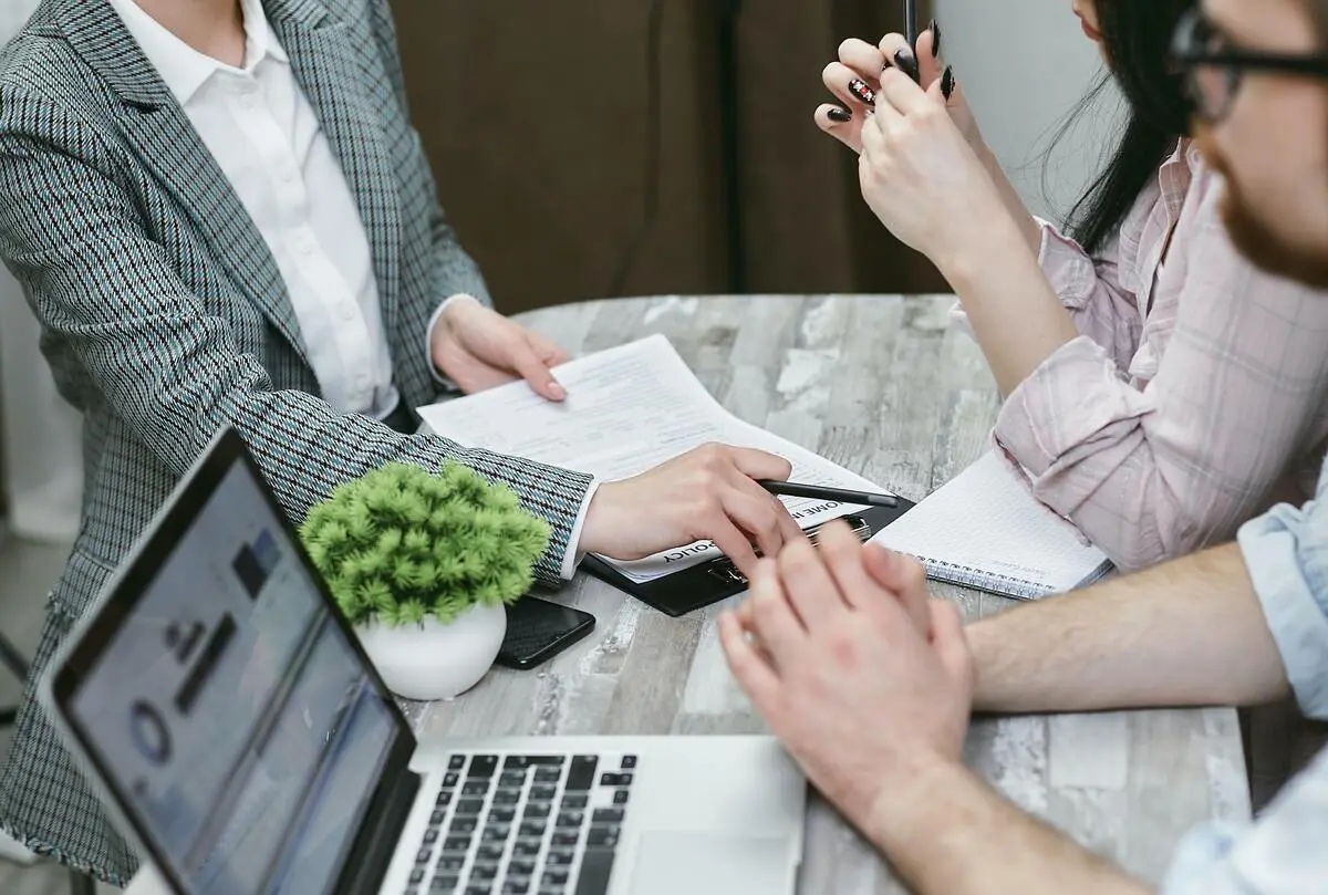 Group of people in a business meeting at a desk with laptops, paperwork, and notebooks.