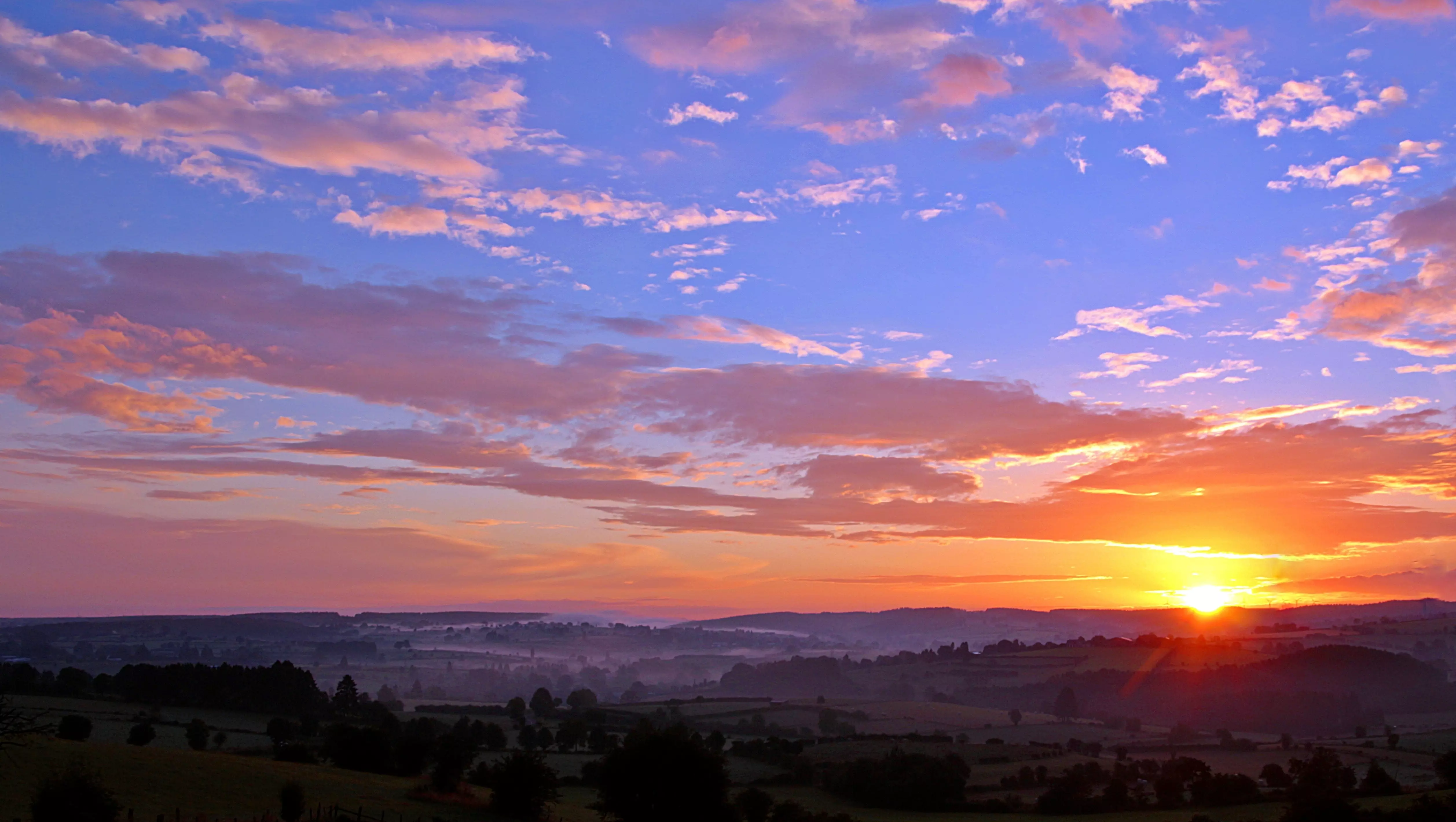Sunrise over rolling hills with colourful clouds in a purple, pink, and orange sky.