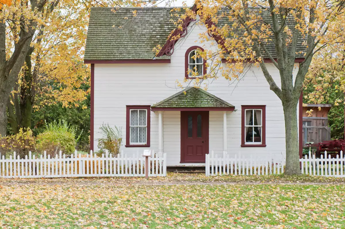 Charming white house with red trim and a small front porch, surrounded by autumn trees and a white picket fence.
