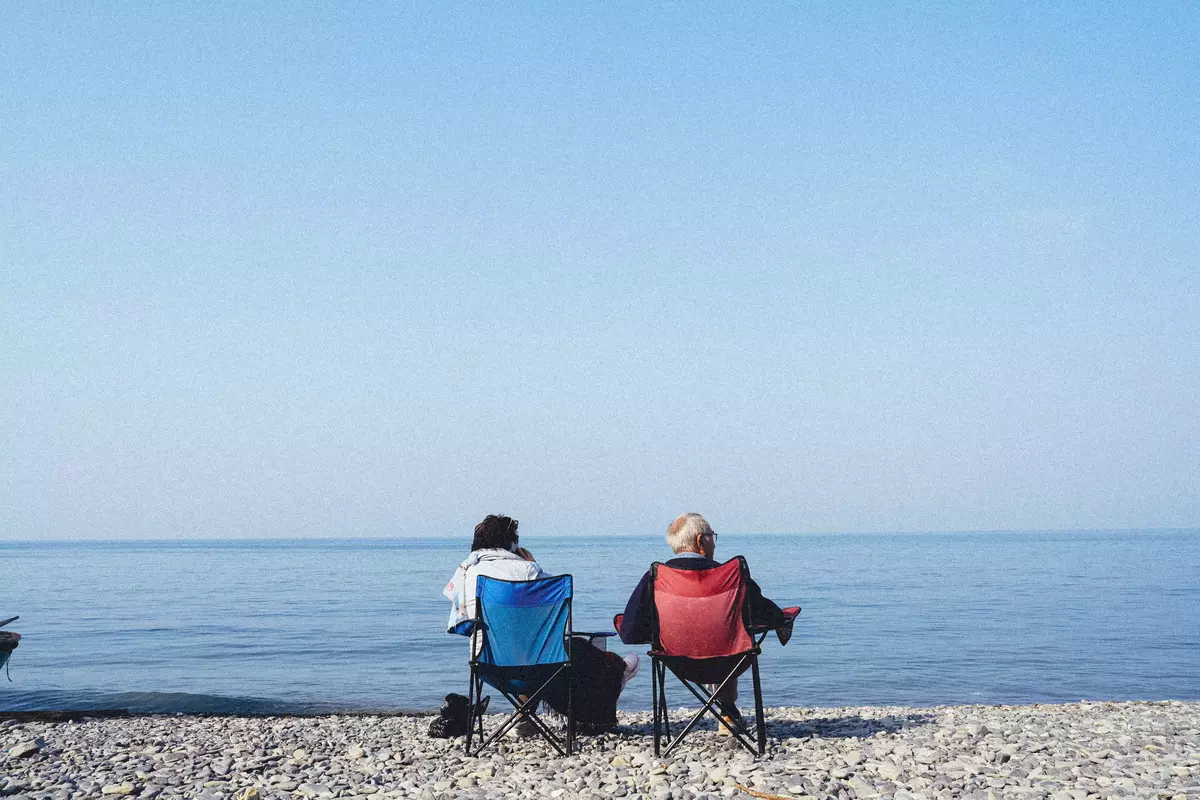 Two people sitting on folding chairs by a calm pebble beach, looking out at the horizon over the sea.