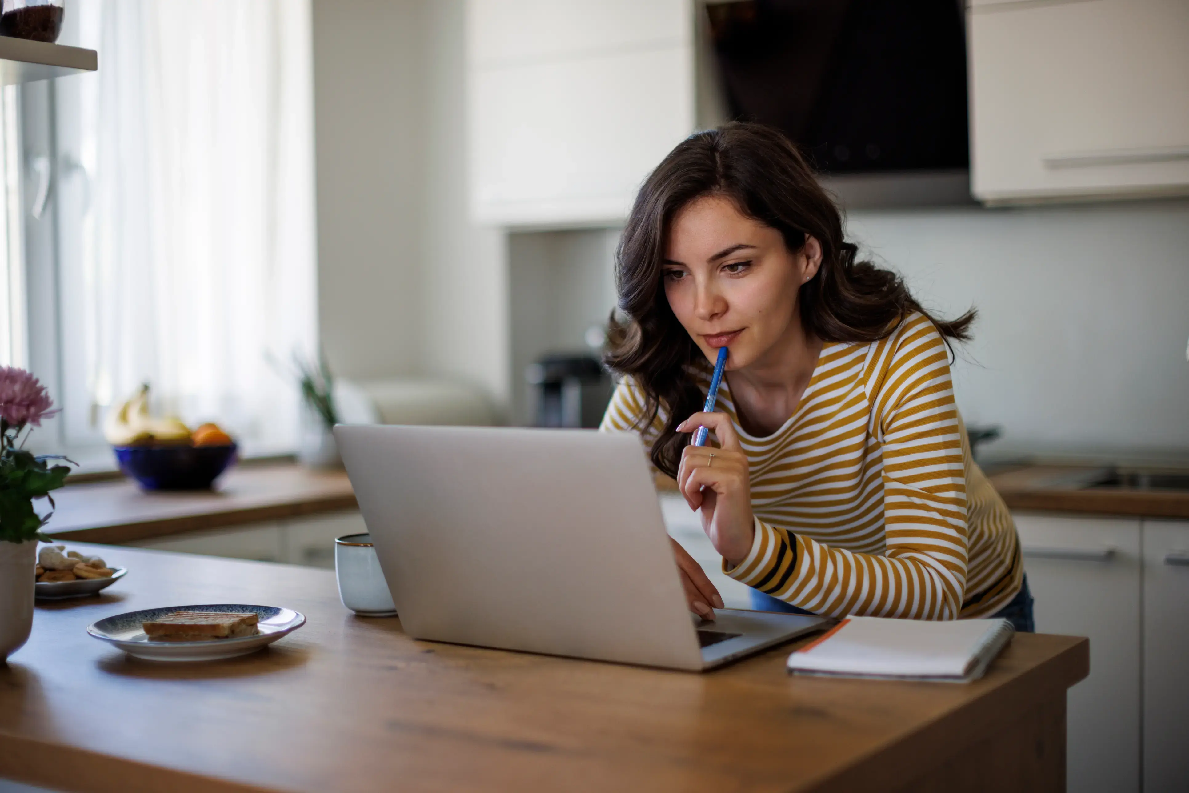 A person sits at a wooden kitchen table working on a laptop, holding a pen thoughtfully near their mouth, with a notebook, coffee cup, and small plate of food nearby in a bright, modern home kitchen.