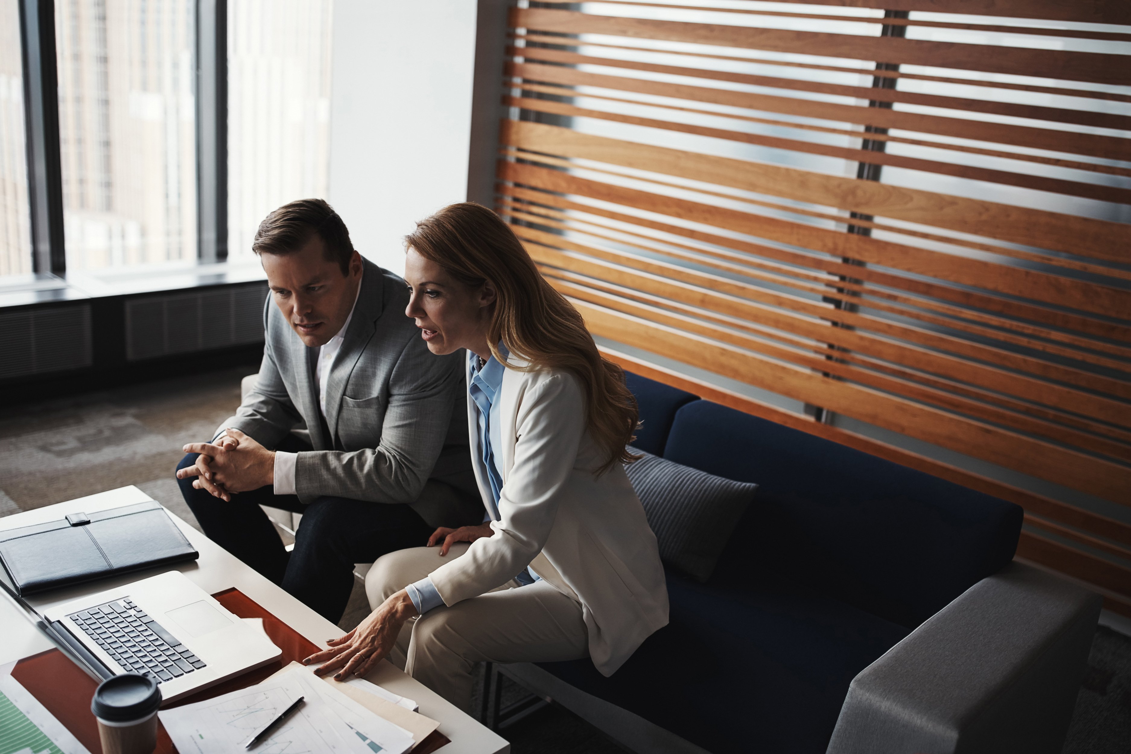 Two professionals seated closely on a modern office couch, leaning forward and looking intently at a laptop placed on a low coffee table in front of them. Their posture and facial expressions suggest focused discussion or problem-solving.