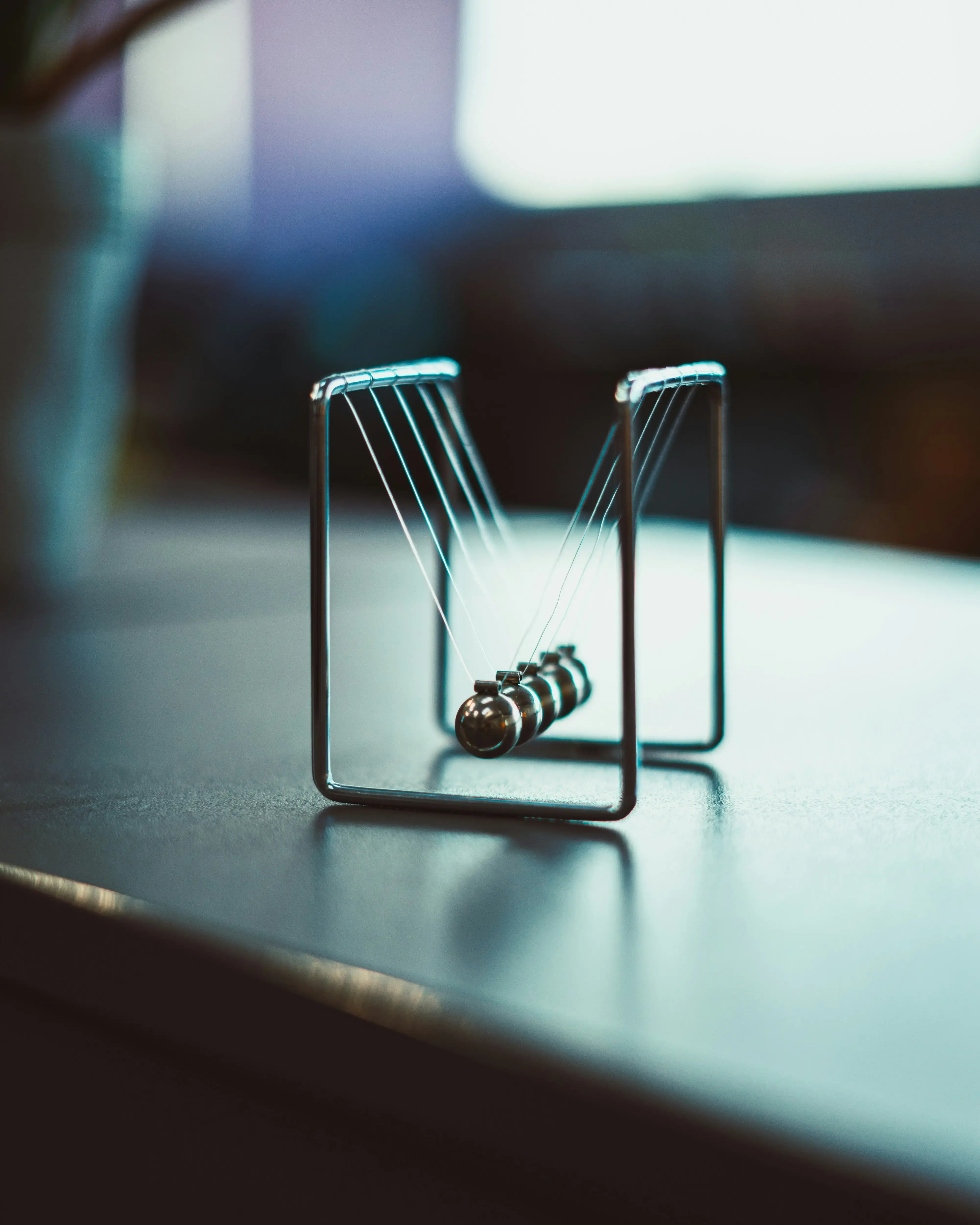 A Newton’s cradle (the desk toy with metal balls that swing and transfer momentum) on a table, with a blurred background and soft lighting.