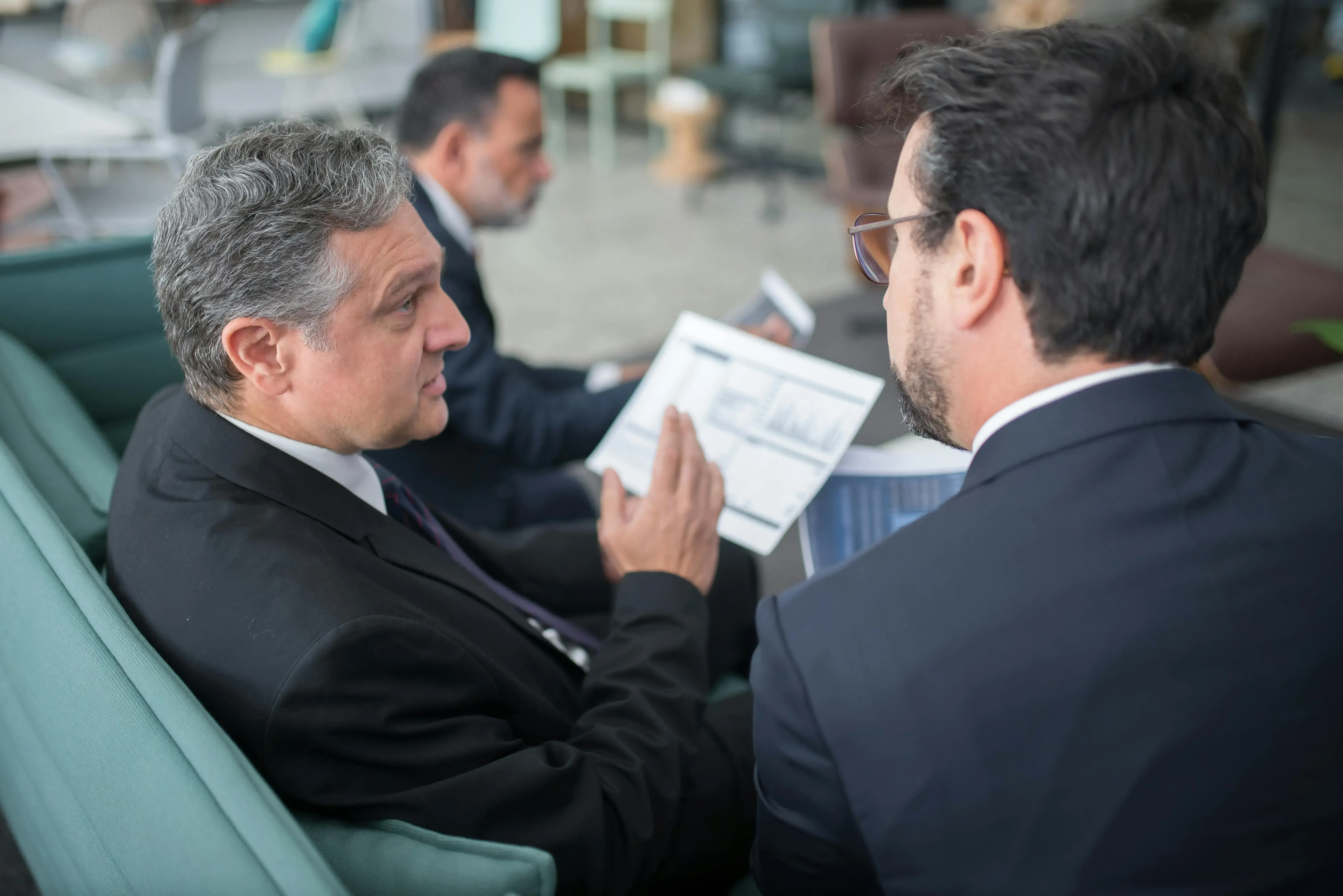 Two businessmen in suits having a discussion while reviewing printed charts and reports, seated on a teal sofa in a modern office setting. Another man in the background is also looking at documents.
