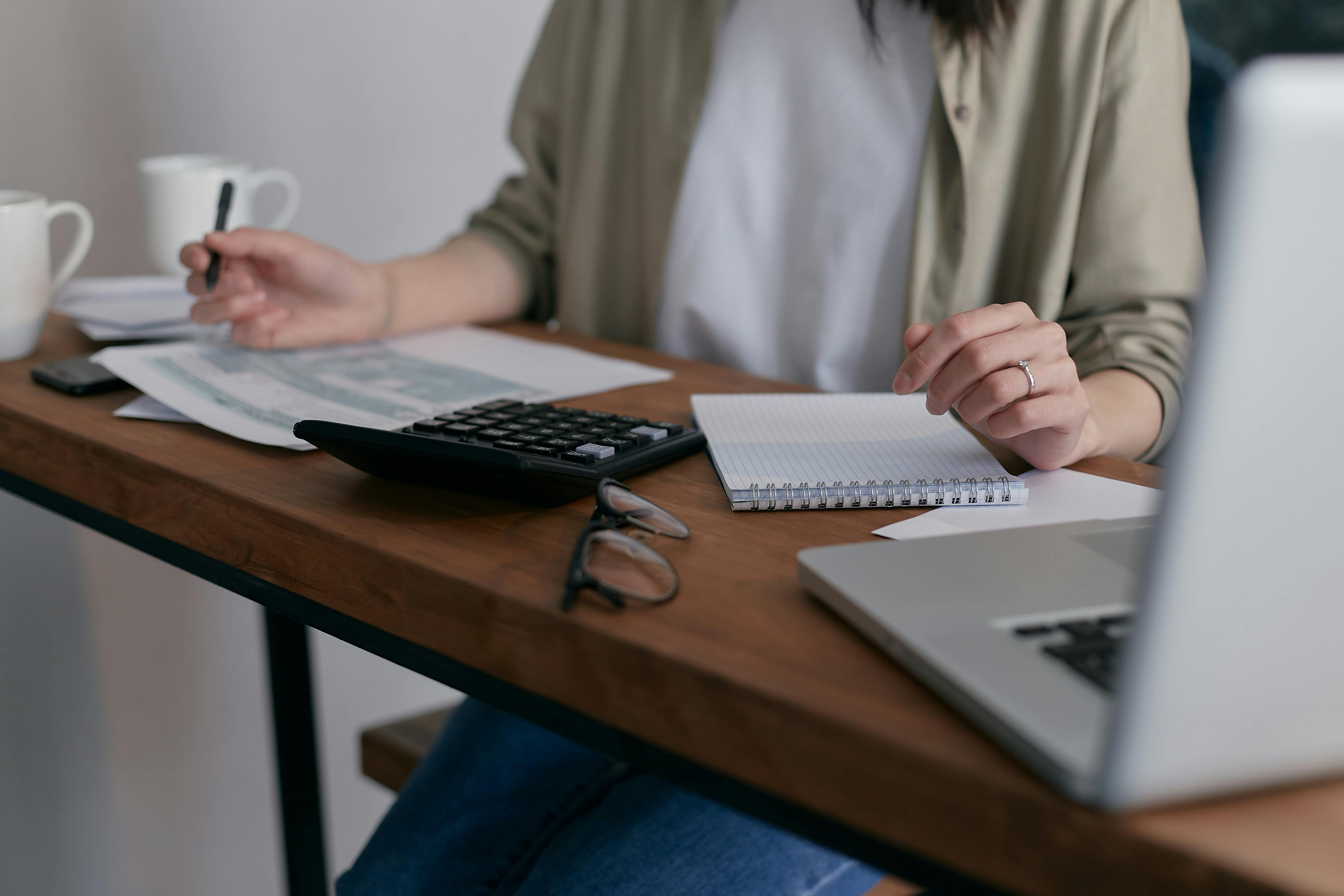 A person sitting at a desk working with financial documents, a calculator, a notebook, and a laptop, suggesting budgeting or accounting work.