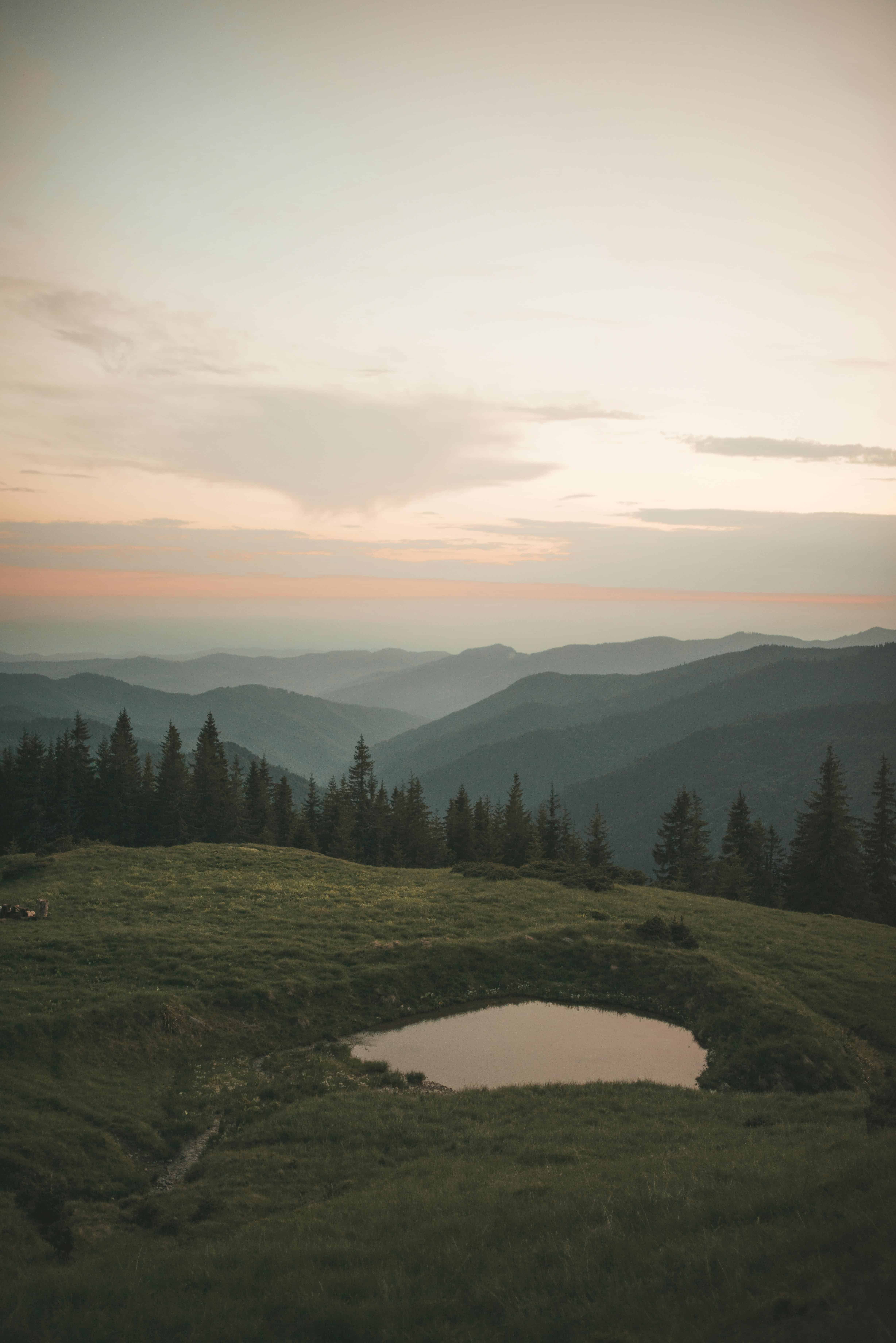 Peaceful mountain landscape at sunset with layers of rolling hills fading into the distance. A small pond sits in the grassy meadow surrounded by evergreen trees, under a soft pastel sky