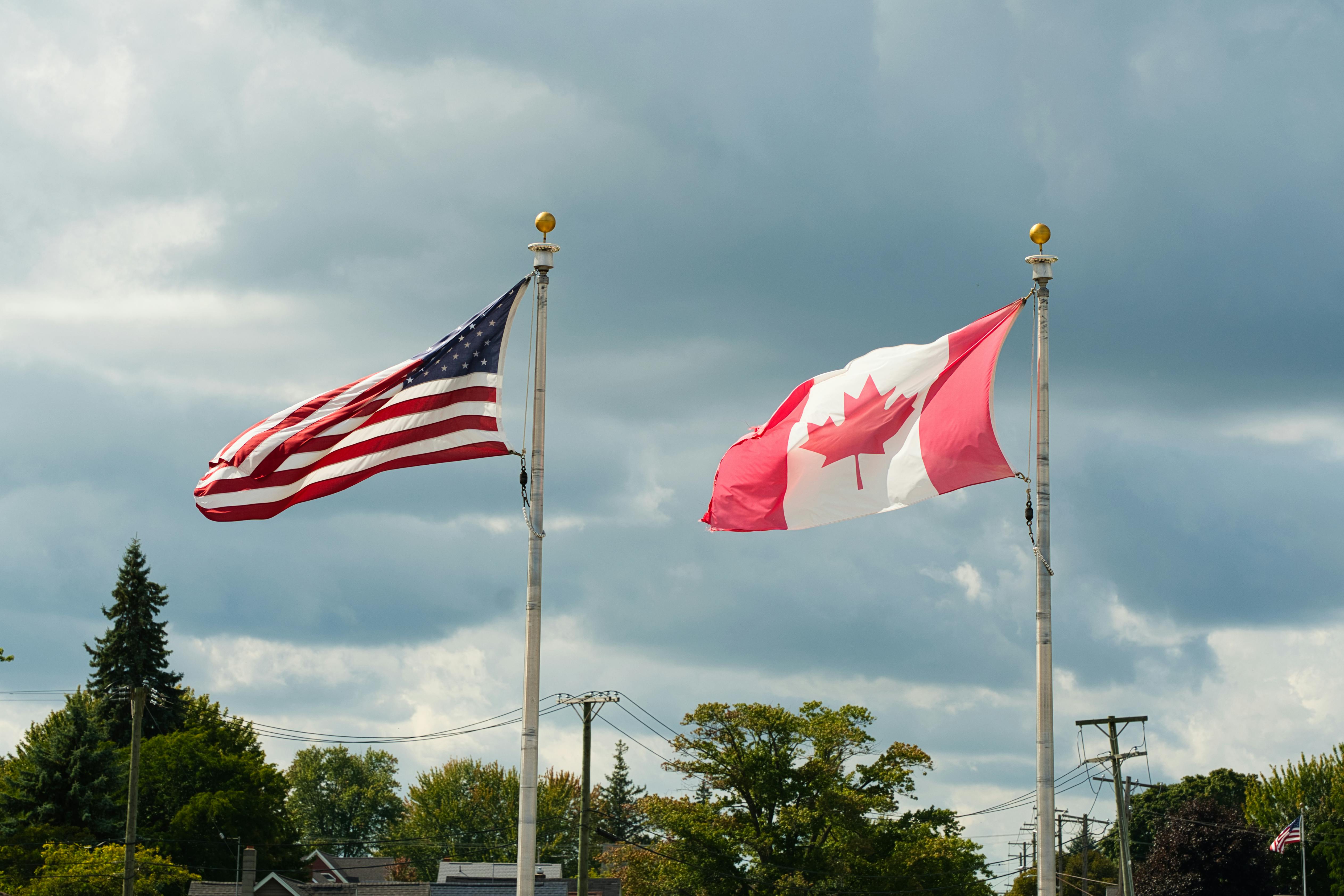 The U.S. flag and the Canadian flag fly side by side on tall flagpoles against a cloudy sky, with trees and rooftops visible below.