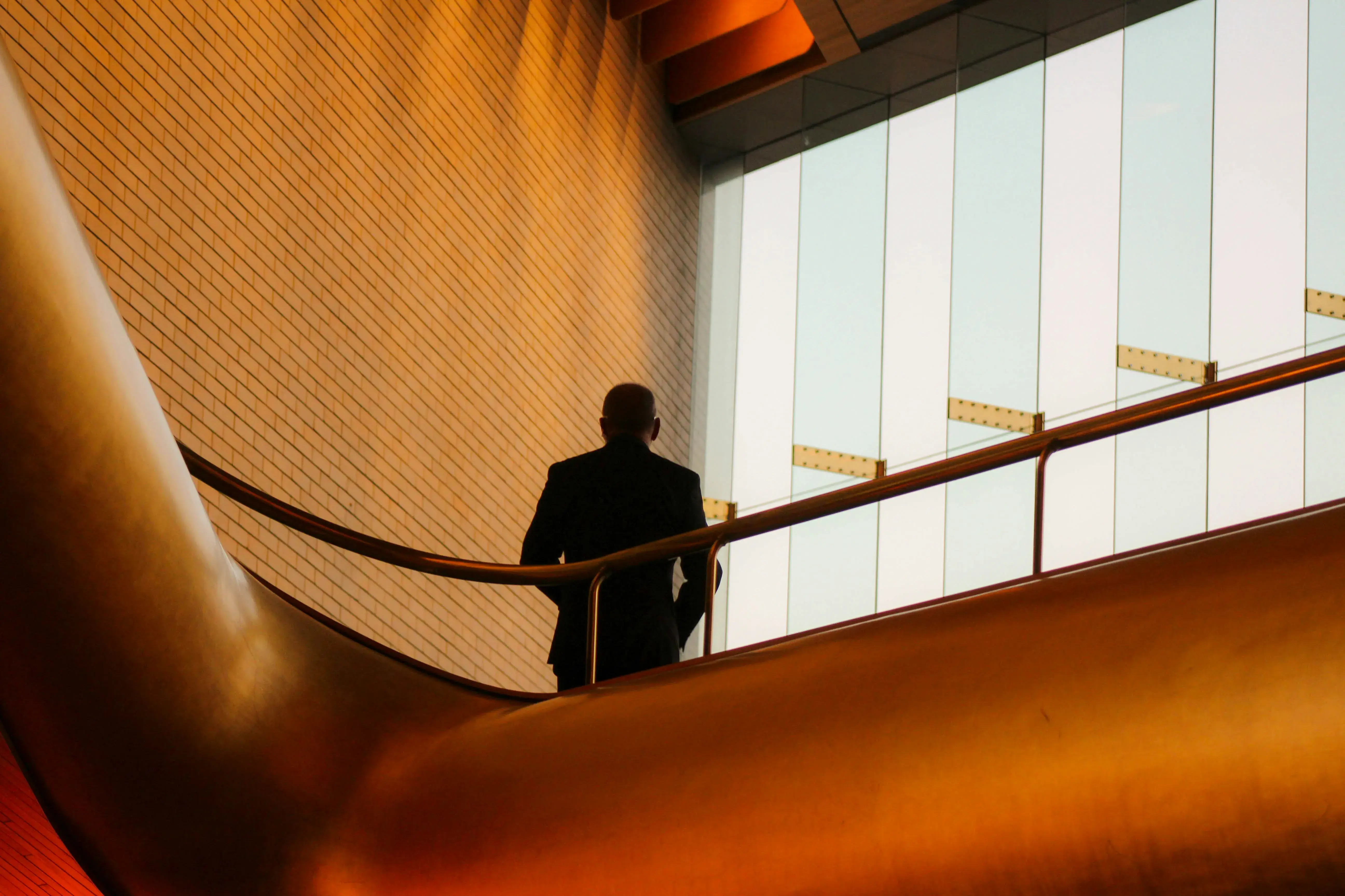 A person in a dark suit walks up a curved staircase inside a modern building, silhouetted against large frosted windows, with warm orange lighting reflecting off the smooth, sweeping architecture.