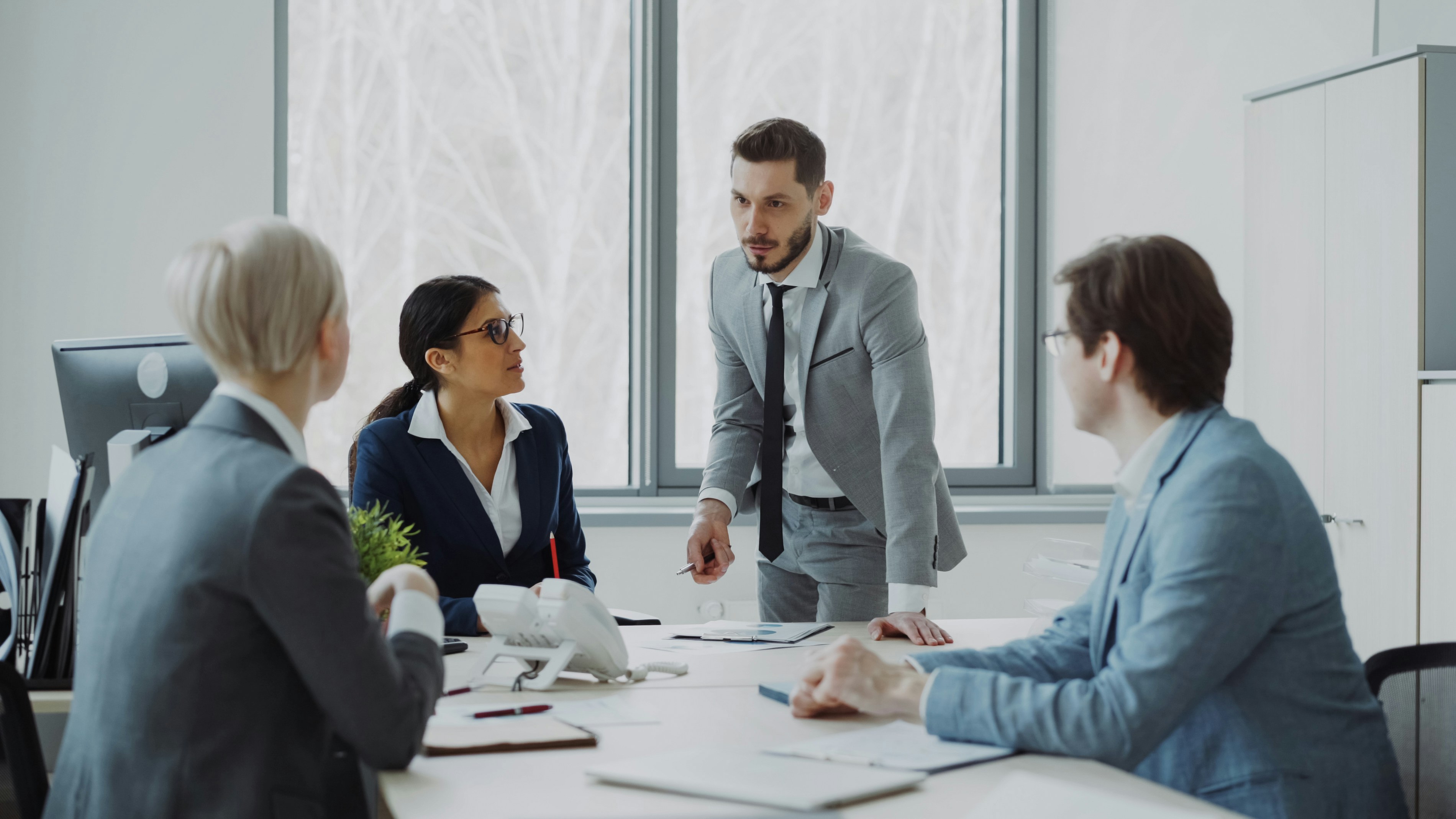 Business team in formal attire having a discussion around a conference table, with one man standing and speaking while others listen attentively in a modern office.
